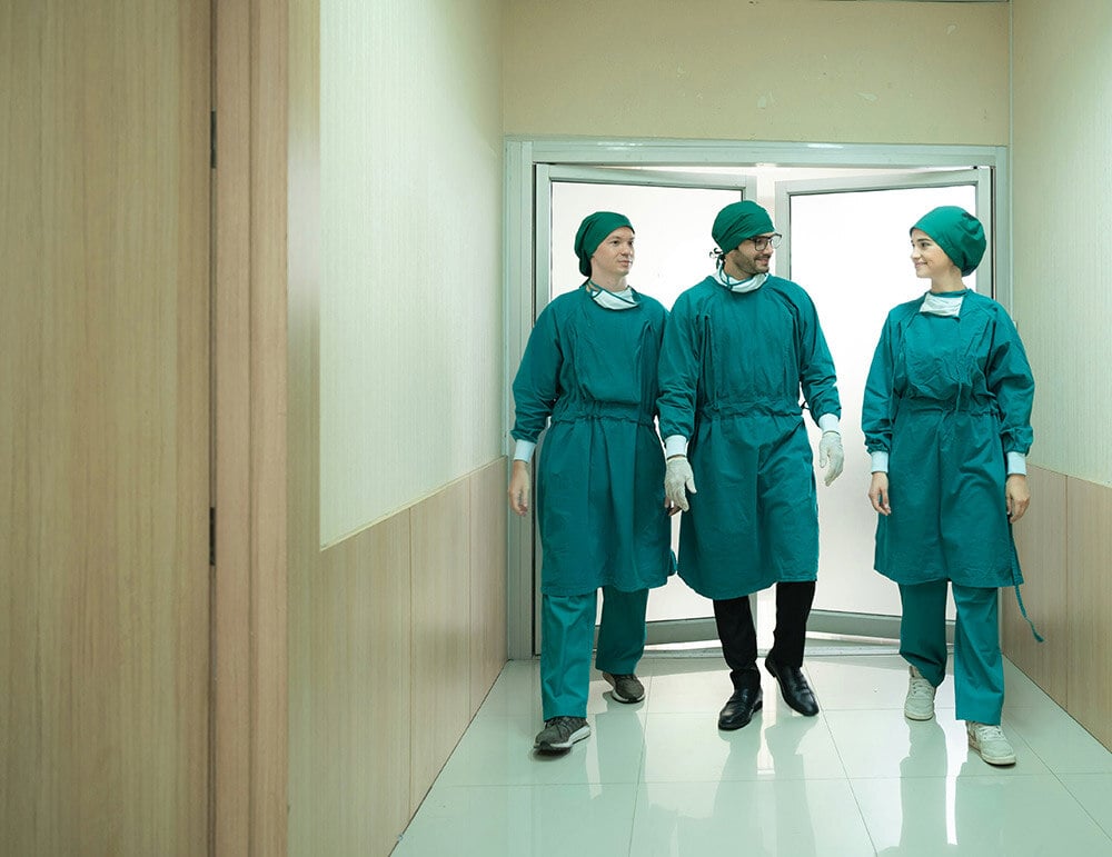 Three medical professionals in green surgical attire walking through a hospital corridor, engaged in conversation.