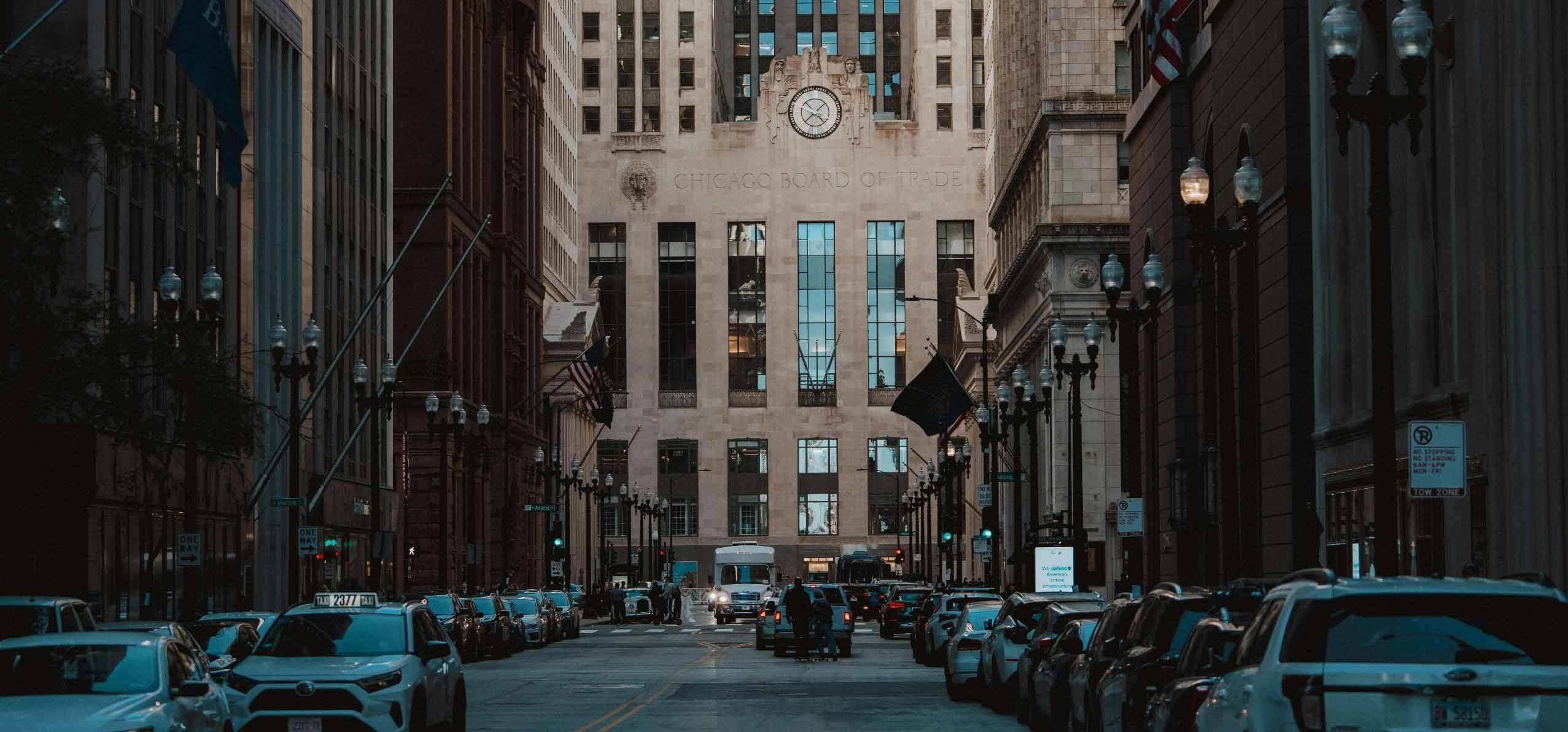 A city street scene in Chicago with parked cars, taxis, and pedestrians, leading to the Chicago Board of Trade building.