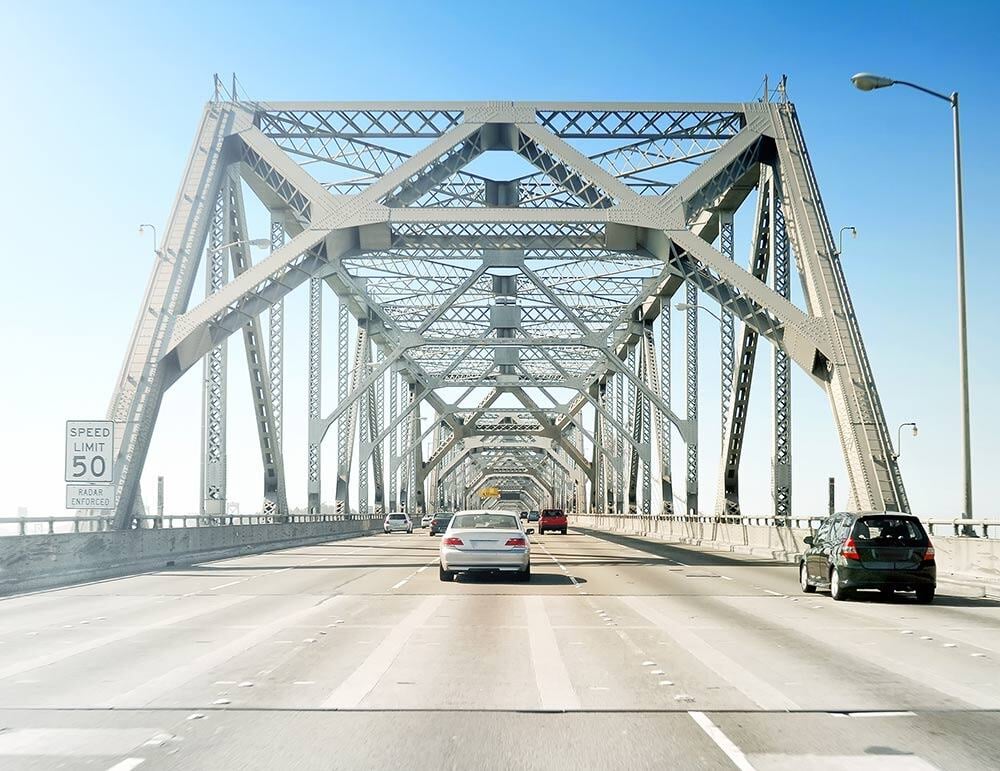 Vehicles driving on a steel truss bridge under a clear blue sky, with a speed limit sign of 50 mph.